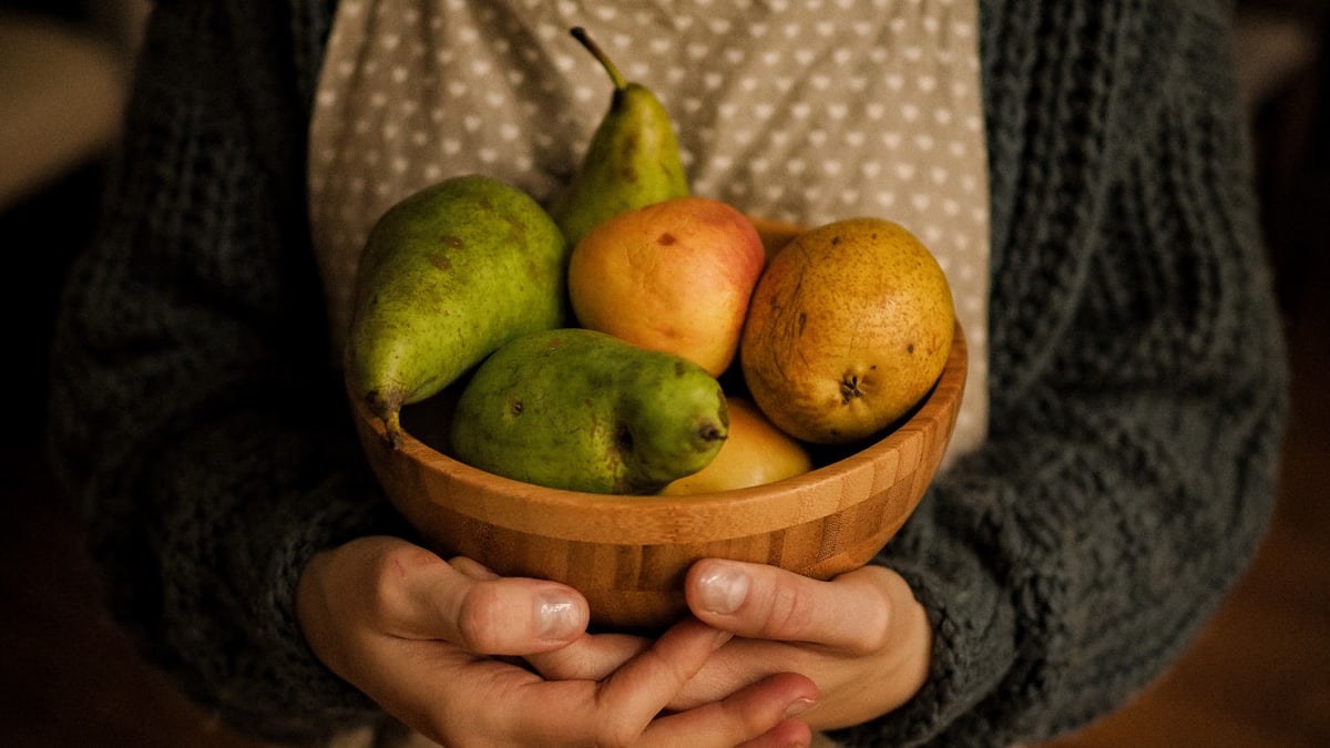 Close-up of hands holding a wooden bowl filled with pears and apples in warm indoor lighting.